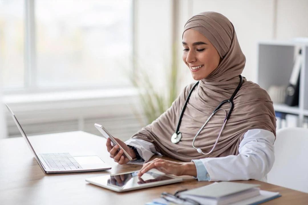 A person with a stethoscope around their neck smiles as they look at a phone in one hand and enter information into a tablet with the other hand.